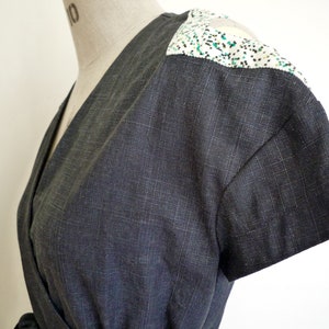 May include: A dark charcoal gray wrap top with short sleeves and a V-neckline. The shoulders feature a contrasting white panel with a speckled pattern of small black and green dots. The fabric has a visible woven texture throughout the garment.