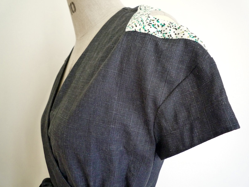 May include: A dark charcoal gray wrap top with short sleeves and a V-neckline. The shoulders feature a contrasting white panel with a speckled pattern of small black and green dots. The fabric has a visible woven texture throughout the garment.