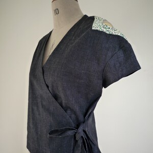 May include: A dark charcoal gray wrap top with short sleeves and a V-neckline. The garment features a tie closure at the side waist and contrasting white shoulder panels patterned with small green and black speckles, displayed on a mannequin.