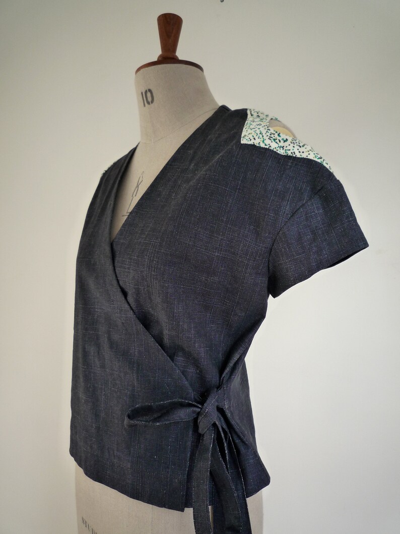 May include: A dark charcoal gray wrap top with short sleeves and a V-neckline. The garment features a tie closure at the side waist and contrasting white shoulder panels patterned with small green and black speckles, displayed on a mannequin.