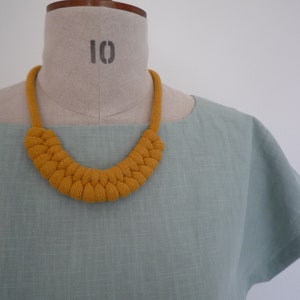 May include: Mustard yellow knitted necklace with a braided design. The necklace is displayed on a mannequin with a light green top. The mannequin has the number 10 printed on it.