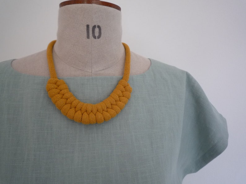 May include: Mustard yellow knitted necklace with a braided design. The necklace is displayed on a mannequin with a light green top. The mannequin has the number 10 printed on it.