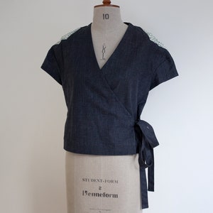 May include: A dark charcoal gray wrap top with short sleeves and a V-neckline. The garment features a side tie closure and light-colored, intricate lace detailing on the shoulders. The fabric has a subtle, fine-lined texture throughout the design.
