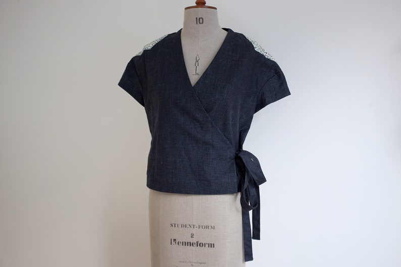 May include: A dark charcoal gray wrap top with short sleeves and a V-neckline. The garment features a side tie closure and light-colored, intricate lace detailing on the shoulders. The fabric has a subtle, fine-lined texture throughout the design.