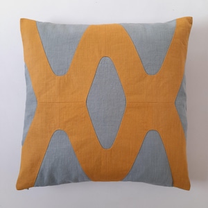 May include: Square decorative pillow with a gray background and a large, geometric, mustard-yellow design. The pillow has a stitched pattern and a soft, textured appearance. The design is symmetrical and modern.