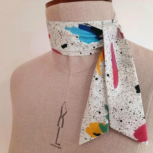 May include: A white neck scarf featuring a black speckled pattern with abstract shapes in blue, yellow, pink, and green. The scarf is tied around the neck of a beige fabric mannequin, with one end hanging down in a pointed shape.