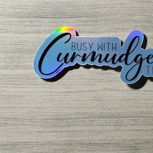 May include: A holographic sticker with the text "Busy with curmudgeonly things" in black lettering.