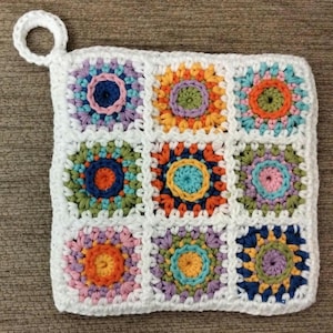 May include: A square, handmade crochet potholder featuring nine colorful floral squares in shades of orange, blue, green, and purple, bordered by white crochet. It has a white loop for hanging. The potholder is approximately 8 inches square.