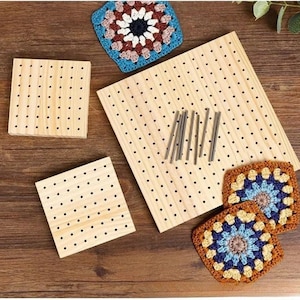 May include: A set of three light-colored wooden blocking boards with evenly spaced holes, accompanied by several thin metal pins. Three colorful, square, crocheted granny squares with blue, white, and brown yarn are positioned near the boards.