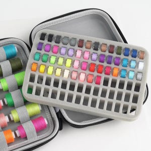 May include: A gray sewing kit with a variety of colorful thread spools. The kit is open, revealing a foam insert with multiple thread spools in various colors, including blue, green, yellow, pink, and red. The kit is designed for organizing sewing supplies.