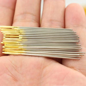 May include: A close-up of a stack of sewing needles. The needles are silver with gold-colored eyes. The needles are held in a person's hand.