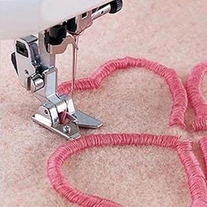 May include: A silver metal sewing machine presser foot is stitching a continuous pink thread pattern onto a light pink felt fabric surface. The needle is positioned within the metal foot, creating a textured, raised heart-shaped design.