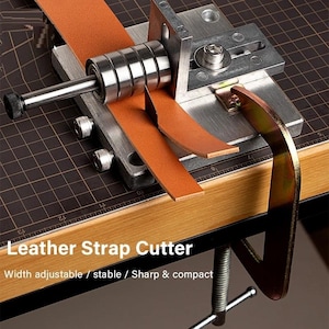 Pode incluir: Um cortador de tiras de couro de metal com uma tira de couro castanho a ser cortada. O texto "Leather Strap Cutter" é visível, juntamente com "Width adjustable / stable / Sharp & compact". O cortador está preso a uma superfície de madeira.