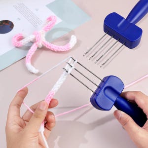 May include: Two blue plastic knitting tools with four metal prongs each, used for creating cord. One tool is actively working on a white and pink variegated yarn cord, while a finished pink and white braided yarn bow lies nearby on the surface.