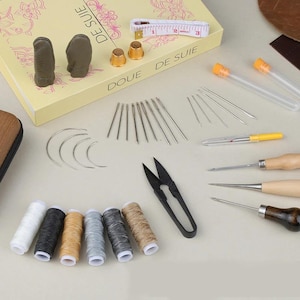 May include: A collection of leathercraft tools, including needles, awls, scissors, thimbles, and spools of thread in various colors. A measuring tape and a box with the text "DOUÉ DE SUITE" are also present.