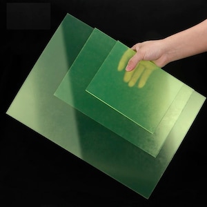 Puede incluir: Una mano sostiene varias láminas rectangulares verdes translúcidas de diferentes tamaños sobre un fondo negro. Las láminas parecen estar hechas de un material flexible, con un ligero brillo. La lámina más grande está en primer plano, con dos láminas más pequeñas detrás.