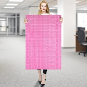 Könnte beinhalten: Eine große, rechteckige, selbstheilende Schneidematte in Pink mit einem weißen Rastermuster, verschiedenen geometrischen Formen und aufgedruckten Winkelmarkierungen. Die Matte ist für Bastelarbeiten und präzise Schneideaufgaben konzipiert.