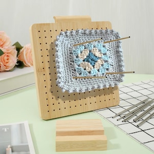 May include: A wooden crochet blocking board with a light blue, beige, and dark blue crocheted square pinned in place. The board has numerous small holes and a wooden handle. Two metal rods are inserted through the crochet square.
