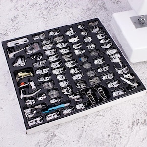 May include: A set of sewing machine presser feet in a black tray with individual compartments. The feet are silver and clear, and the tray is inside a white box. A white sewing machine is partially visible in the background.