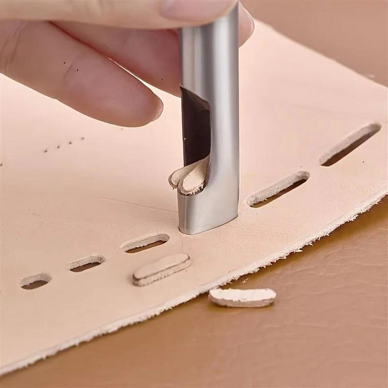 May include: A metal leather punch tool cutting a series of oval holes in a piece of light tan leather. The tool is silver and has a curved cutting edge. The leather is on a brown surface.