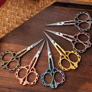 May include: An assortment of decorative embroidery scissors in various colors, including gold, bronze, and iridescent blue. The scissors have ornate handle designs and sharp, pointed blades. They are arranged on a dark wooden surface.