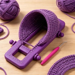 May include: A rectangular purple plastic knitting loom with a partially completed purple yarn project attached to the pegs. A pink handled metal hook tool lies nearby, accompanied by two balls of purple yarn and a separate piece of purple fabric.