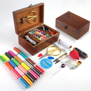 May include: A wooden sewing box with an open lid, revealing spools of colorful thread, scissors, and other sewing supplies. Additional items include a closed wooden box, spools of thread, and a strawberry-shaped pin cushion.