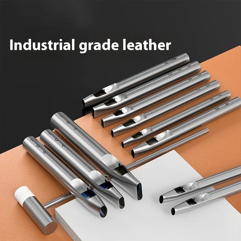 May include: A collection of silver-colored leather hole punches of various sizes, arranged on a white and brown surface. The tools have cylindrical bodies with numbered markings and rectangular or circular cutting ends. The text "Industrial grade leather" is displayed at the top.