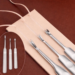 May include: A collection of silver leathercraft tools, including stitching groovers and pricking irons, displayed on a piece of tan leather. The tools have various prong configurations for creating precise stitching holes. The background is a warm brown.