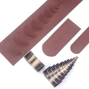 May include: A collection of leather crafting tools, including strips of brown leather with crescent-shaped cutouts, and metal cutting tools. The tools are arranged on a white surface, showcasing the variety of shapes and sizes available for leatherwork projects.