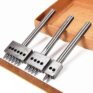 Puede incluir: Tres herramientas metálicas para trabajar el cuero con diferentes números de púas y agujeros. Las herramientas tienen mangos plateados y cabezas rectangulares. Se ve una tira de cuero marrón con agujeros perforados, junto con una caja de cartón en el fondo.