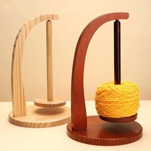 May include: Two wooden yarn holders. One is natural wood, the other is stained dark brown. The brown holder has a yellow yarn ball on it. Both have a curved arm and a central dowel for holding yarn.