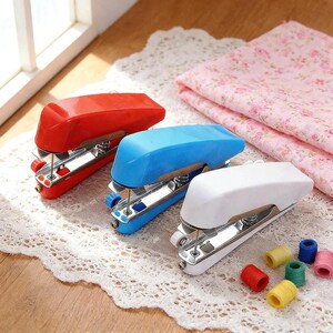 May include: Three handheld sewing machines in red, blue, and white with metallic silver components. Each device features a plastic casing, a metal presser foot, and a needle mechanism. Several small, colourful thread spools are scattered nearby.