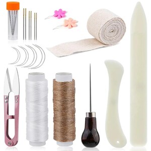 May include: A collection of leathercraft tools and supplies, including needles, thread, scissors, a bone folder, and a stitching awl. The kit also includes a roll of beige fabric tape and two flower-shaped pins. The items are arranged on a white background.