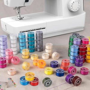 May include: A white sewing machine with a variety of colorful thread spools and bobbins. The spools are stacked in columns and scattered around the machine, showcasing a range of colors including pink, blue, yellow, and green.