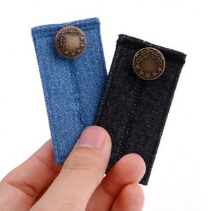 May include: Two rectangular denim fabric waist extenders, one in light blue and one in black, each featuring a centred metallic bronze-toned button embossed with a circular leaf and star pattern, designed to provide additional room for clothing.