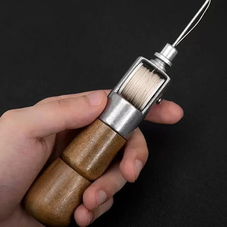 May include: A leather stitching awl with a wooden handle and a silver-colored metal body. The awl has a spool of white thread and a curved needle, designed for hand sewing leather and other materials.