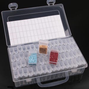 May include: A clear plastic storage box with a hinged lid and handle. The box is divided into numerous small compartments, each designed to hold small items. Three small plastic bags filled with colorful beads are inside the box.