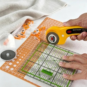Può includere: Un taglierino rotativo giallo con lama da 45 mm, un righello quadrato trasparente per quilting con segni verdi, un tappetino da taglio arancione con linee bianche e diverse clip da cucito arancioni su tessuto bianco.