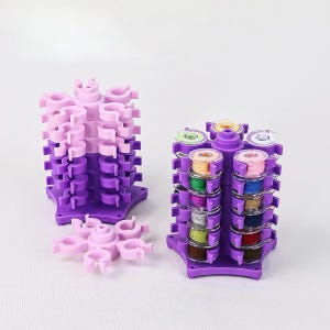 Op de afbeelding: Twee paarse en roze garenspoelorganizers. De ene is leeg, de andere bevat spoelen met kleurrijk garen. De organizers hebben een stervormige basis en meerdere niveaus voor het vasthouden van spoelen. Een losgemaakt roze stuk staat op de voorgrond.