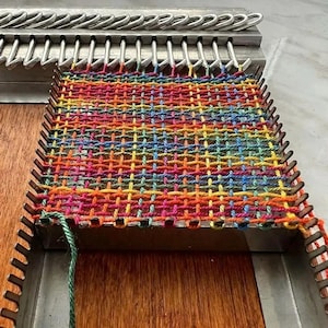 May include: A metal weaving loom featuring a grid of silver prongs holding a vibrant, multi-colored yarn pattern. The woven fabric displays a complex crisscross design in shades of red, orange, yellow, green, blue, and purple, secured by the metal frame.