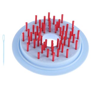 May include: A light blue circular knitting loom with numerous vertical red pegs arranged in a ring pattern, featuring a central opening and a separate light blue plastic yarn needle positioned to the left of the main loom structure.