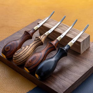 Puede incluir: Cuatro herramientas de marroquinería con mangos de madera y hojas de metal se exhiben en una bandeja de madera. Los mangos varían en color, desde marrón oscuro hasta negro, y están numerados del 1 al 4. Las hojas son plateadas con detalles dorados.