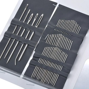 May include: A collection of sewing needles in various sizes and shapes, neatly arranged in a black fabric case. The needles are silver-colored and appear to be made of metal. The case is open, displaying the needles in organized rows.