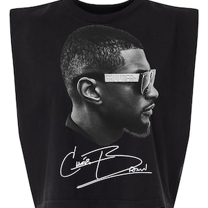 May include: A black sleeveless t-shirt with padded shoulders featuring a high-contrast black and white profile portrait of a person wearing sunglasses with the text GAULTIER on the frame, and a white signature graphic reading Chris Brown below.