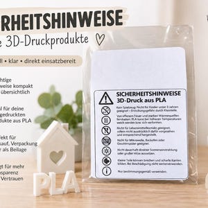 Op de afbeelding: Een doorzichtige plastic zak met een witte rechthoekige veiligheidsinformatiekaart voor 3D-geprinte PLA-producten. De kaart bevat een waarschuwingsdriehoek, zes ronde iconen en Duitse tekst met veiligheidsinstructies en waarschuwingen.