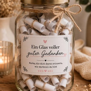 Op de afbeelding: Een doorzichtige glazen pot met een gouden deksel, gevuld met talloze kleine witte papierrolletjes vastgebonden met touw. Een wit etiket op de voorzijde bevat de Duitse tekst: Ein Glas voller guter Gedanken, Worte, die dich daran erinnern, wie wertvoll du bist.