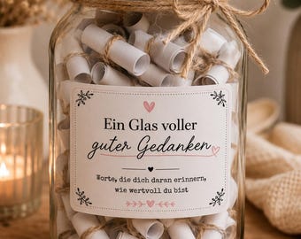 Puede incluir: Un frasco de vidrio transparente con tapa dorada, lleno de numerosos pequeños rollos de papel blanco atados con cordel. Una etiqueta blanca en el frente presenta el texto en alemán: Ein Glas voller guter Gedanken, Worte, die dich daran erinnern, wie wertvoll du bist.