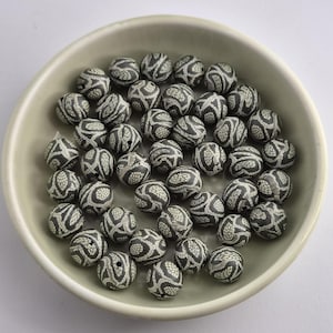 May include: A collection of spherical beads in a light-coloured bowl. Each bead features a dark grey and off-white patterned surface with a repeating, textured, scale-like design. The beads are round, uniform in size, and have a small hole.