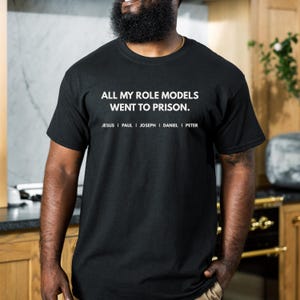May include: Black t-shirt with the text "ALL MY ROLE MODELS WENT TO PRISON." and the names Jesus, Paul, Joseph, Daniel, and Peter printed below the text in white. The shirt is made of a soft material.